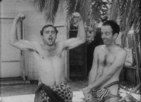 A black-and-white film still of two shirtless men wearing material resembling loincloths below the waist. The man in the left has both arms raised, elbows bent at 90 degrees, flexing to show bicep muscles; his mouth is open. The man on the right is looking away from him, smiling, his arms hanging in front of him, left hand grabbing right wrist.