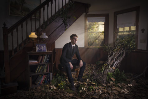 A man with glasses wearing a black suit is seated on a stool in the middle of a living room in an old house looking into the distance. A wood staircase is behind him, and a three-shelf bookshelf with books is to his right. The floor is covered in fallen leaves and moss, and a tree grows through the center of the floor, growing out of the room through an open window to the man's left.
