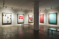 A part of the seventh floor gallery of The Andy Warhol Museum. The gallery has white walls, a pillar right of center, and a bench on the right. There are three large, rectangular paintings by Andy Warhol on the left-hand wall and two on the right-hand wall. The first artwork on the left is black and white and depicts black and white feet that show the steps of a dance. The middle one is an outline of a Cambell's Soup can with only the red of the soup label painted. The next one is a Cambell's Soup can with the label peeling off. The red and yellow of the soup label is painted and the can is painted grap an black. On the right-hand wall, the painting on the left is red paint on a white canvas in a pattern over and over. The painting on the right is painted almost entirely a teal color with some small patterns in the top-left corner.