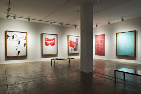 A part of the seventh floor gallery of The Andy Warhol Museum. The gallery has white walls, a pillar right of center, and a bench on the right. There are three large, rectangular paintings by Andy Warhol on the left-hand wall and two on the right-hand wall. The first artwork on the left is black and white and depicts black and white feet that show the steps of a dance. The middle one is an outline of a Cambell's Soup can with only the red of the soup label painted. The next one is a Cambell's Soup can with the label peeling off. The red and yellow of the soup label is painted and the can is painted grap an black. On the right-hand wall, the painting on the left is red paint on a white canvas in a pattern over and over. The painting on the right is painted almost entirely a teal color with some small patterns in the top-left corner.