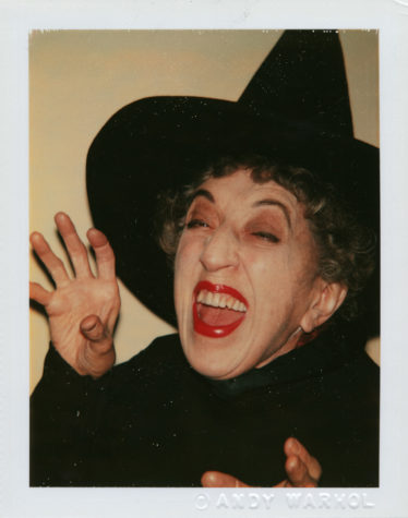 Photograph of a woman wearing a black pointed witch hat and a black cape. Her hand is raised by her face in a claw shape, and mouth looks like she is cackling.