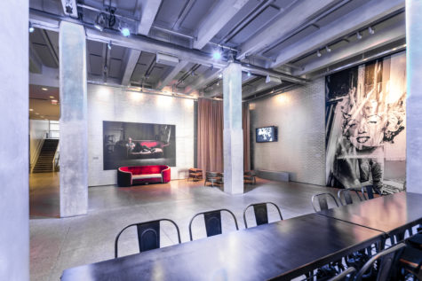 A large room with high ceilings with a brick accent wall painted silver. This space includes a red sofa, tables, chairs and photographs on the walls featuring Andy Warhol.