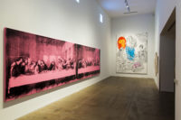 A section of the fourth floor gallery of The Andy Warhol Museum. The gallery has white walls. On the left wall is an artwork by Andy Warhol depicting the last supper in red tint. On the wall straight ahead is another artwork of Andy Warhol's on white canvas that has black outlines of four angels, the one in the middle had the clothing outline filled in with light blue paint. The angel at the bottom, who is laying on the ground, is holding a child that is painted pink.