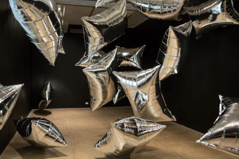 A gallery on the fifth floor of The Andy Warhol Museum that has black walls. In the gallery are many floating large, rectangular silver balloons.