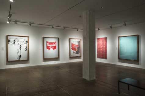 A part of the seventh floor gallery of The Andy Warhol Museum. The gallery has white walls, a pillar right of center, and a bench on the right. There are three large, rectangular paintings by Andy Warhol on the left-hand wall and two on the right-hand wall. The first artwork on the left is black and white and depicts black and white feet that show the steps of a dance. The middle one is an outline of a Cambell's Soup can with only the red of the soup label painted. The next one is a Cambell's Soup can with the label peeling off. The red and yellow of the soup label is painted and the can is painted grap an black. On the right-hand wall, the painting on the left is red paint on a white canvas in a pattern over and over. The painting on the right is painted almost entirely a teal color with some small patterns in the top-left corner.