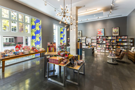 A room with gray walls, white ceiling and a gray floor. Large windows to the outside on the left side wall. There is a white pillar in the middle of the room. Tables with various merchandise are scattered throughout the space. There are shelves lining the back and right side wall with books and other merchandise on them. Two chairs are on the floor on the right side. There are lights hanging from the wall in the middle that are metal with spokes coming out and small lights at the end.