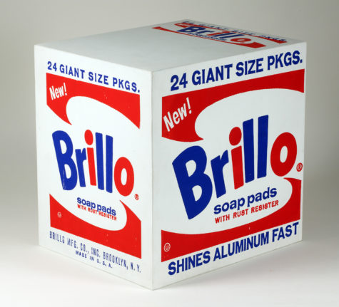 A box with only two sides and the top showing. "24 Giant Size Pkgs." is written along the top of the right and left sides in blue and "Shines Aluminum Fast" is written along the bottom of the right in blue. On the left side on the bottom, "Brillo Mfg. Co. Inc. Brooklyn, N.Y. Made in the U.S.A is written on the bottom in blue. The Brillo soap pads logo is painted in the middle of the two sides and a smaller version of the logo is painted on the right side of the top of the box. The logo has the b, r and both l's in blue and the i and the o in red. "soap pads" is underneath "Brillo" in blue. "With Rust Register" is under "soap pads" in red. Surrounding the words is a red border on top and bottom edges only that has "New!" painted on the top-left side in white.