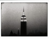 Still from the "Empire" film of the Empire State Building in New York City in black and white. The top of the building is showing, with the sky in the background. Silhouettes of the tops of other buildings can be seen at the bottom of the frame.