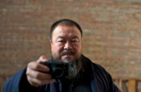 Artist Ai Wei Wei, an asian man with a beard that is just beginning to turn gray, stands against a faded red brick wall, right arm extended toward the viewer, taking a selfie.