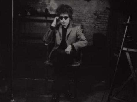 A black and white film still from Bob Dylan's screen test in which the musical icon sits in a chair wearing dark sunglasses and a blazer.