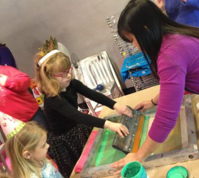 A young woman in a purple sweater with long brown hair helps a little girl with blonde hair and pink glasses drag green ink across a screen to produce a print.