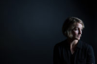 A woman with short blonde hair sits in the lower right of the image. The background is dark, almost black, and she wears a black shirt. She is looking away from the viewer and the side of her face away from the viewer is illuminated.
