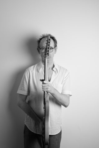 A black and white image with Arto Lindsay holding an electric guitar in front of him, the head of the guitar is in front of his face and turned sideways, the body of the guitar is in front of his torso.