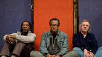 Three men, members of Matthew Shipp Trio, sitting on the ground in front of a wall with three vertical panels of color, blue, red, and blue.