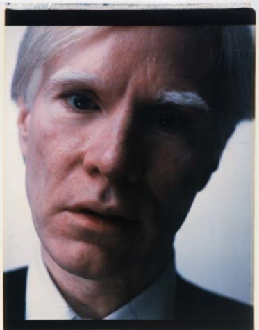 A close-up photograph of Andy Warhol's face, tilted slight to the right side of the screen, against a plain white background.
