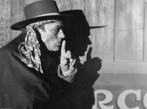 a black and white image of a man in a hat with a finger raised to his lips as if he is gesturing for someone to be quiet. He faces right, and his shadow falls crisply on the wall behind him.