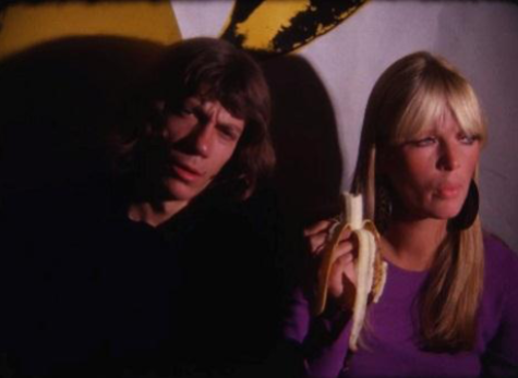 A film still featuring a man with short brown hair and one with long blonde hair, who is wearing a purple shirt and holding a peeled banana.