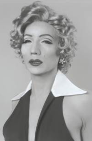 A black and white photo of a man dressed in drag as Marilyn Monroe, complete with curly blonde wig, bold lip, and beauty mark.