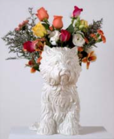 A white ceramic sculpture of a small, seated terrier dog with fresh flowers sprouting from behind its head, as if it is a vase.