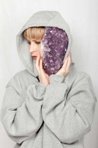 A young woman with blonde hair tucked into the raised hood of her light grey sweatshirt covers her face with a large purple crystal.