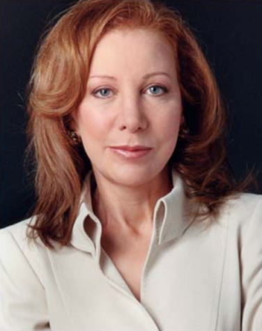 A woman with red hair in a white button up smiles at the camera against a black background in this professional photograph.