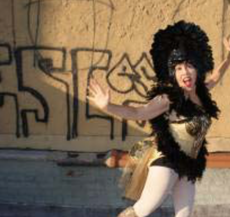 A woman in a black and gold show girl costume dances, arms outstretched, in front of a graffiti covered wall.