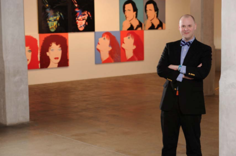 Eric C. Shiner stands in a gallery that contains several of warhol's iconic screen printed portraits wearing a black suit and striped bowtie, his arms folded over his chest.