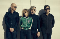 A group of four people, standing left to right in front of a wall: a bald man wearing a black shirt, blue jeans and dark sunglasses; a woman with curly blonde hair, wearing a green jacket, blue jeans and dark sunglasses; a man with salt and pepper hair wearing a black shirt, blue jeans and dark sunglasses; a man with brown hair wearing a black shirt, blue jeans and dark sunglasses.