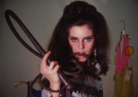 A film still depicting a young woman with dark brown hair holding a whip in her teeth and gazing intensely into the camera.