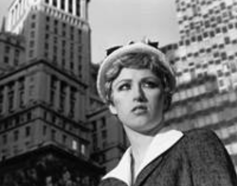 A low angle black and white photograph of a young woman's concerned face against the backdrop of a cityscape.