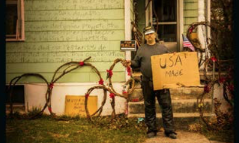 A photograph of a man standing in front of a house with several wreaths behind him holding a cardboard sign that reads USA Made.