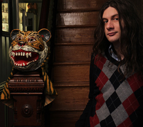 A young man with long black hair in a red and grey argyle sweater stands in front of a deep brown staircase with a tiger's head carved into the bannister.