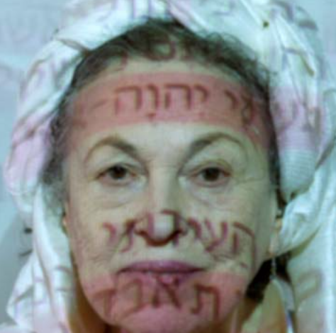 A woman with her head covered in white fabric stares toward the camera, red characters projected over her face.