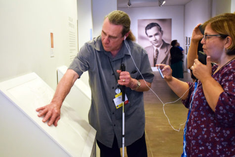 A man with long hair tied back in a ponytail who is holding a cane in one hand reaches out to touch a white plastic tactile reproduction of one of Andy Warhol’s works. He and a woman with short brown hair share a pair of earbuds attached to an audio guide device.