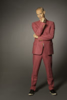 A man with short, gray, thinning hair and a thin mustache is standing wearing a pink suit with gold buttons and black sneakers. He has his left arm under his right elbow and his right thumb and index finger are wrapped around his chin. He's looking at the camera.