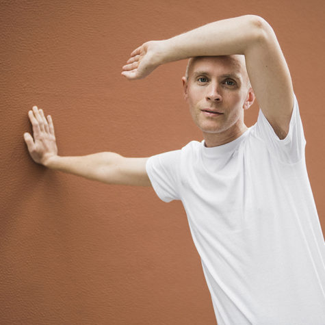 A man in a bright white t-shirt stands to the right of the image, staring directly at the viewer. He is propped up against the muted orange wall to his side. His right arm is outstretched as he pushes against the wall with the palm of his hand. His left arm is bent in a ninety-degree angle, with his hand pointing towards the wall. His forearm rests on the top of his head, casting a slight shadow above his eyes.