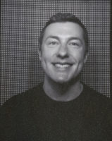 A black and white photo of a man with short hair and wearing a black shirt, taken from the chest up. He is smiling and looking slightly off-camera. The background is comprised of small black squares with white lines around each square.