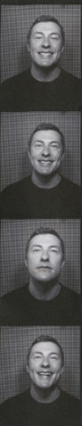 A black and white photo strip of four photos of a man with short hair and wearing a black shirt, taken from the chest up. The background is comprised of small black squares with white lines around each square.