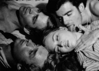 A black and white film still of three men and a woman lying on a surface. Only their heads down to their chests are in the frame. The men on the left have short hair and are looking up towards the ceiling. One man is wearing a button down shirt. The other is shirtless. On the right side of the frame, the other man, who has short, dark hair and is wearing a necklace, is pressing the neck and licking the face of the woman laying in front of him. The woman has light hair and her eyes are closed.