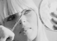 A black and white film still of a closeup of a woman with blonde hair. She has scissors in her hand and is cutting her bangs of her hair while looking into a pocket mirror that she is holding.