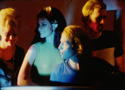 A film still of two women and one man. The image captures them from the waist or chest up. The woman on the left of the frame has short, blonde hair and is looking down and smiling. She's wearing a blue shirt and a pearl necklace. The woman next to her has long, dark hair with daffodils pinned in various places throughout, and is wearing a green shirt. She is looking at the woman in the blue shirt and smiling. The man in front of the woman with the green shirt has long, blonde hair and is wearing a blue t-shirt. He is looking at the woman with the blue shirt on. The man in the right of the frame has short, blonde hair and a black and white t-shirt on. He is looking off to the right of the frame.