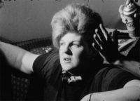 A black and white film still of a woman looking off to the left of the frame. She is sitting on what seems to be a floor in front of a bed. Her arms are up on the bed. Someone off the right of the frame is spraying hairspray into her short, light-colored hair.