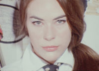 A film still of a woman with long, red hair and wearing a white shirt with a black and white polka dot tie. She is looking straight at the camera.