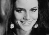 A black and white film still of a closeup of a woman with long, dark hair, smiling and looking a little to the left of the camera. She has dangling earrings on that are balls with polka dots.