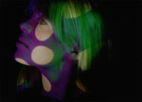 A film still of a closeup of a woman's face. She is looking towards the left of the frame and has her chin up. She has blonde hair and green, purple and white colored lights are being projected onto her face. The background is black.
