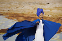 A woman wearing a white dress, blue cape, and blue head wrap is standing sideways towards the camera. She is looking at the camera with her right arm pointed towards the camera and her left arm pointed towards the right side of the image. Her cape is blowing in the wind behind her. She is wearing dark, red lipstick and blue eye shadow. She also has a series of white dots going down her forehead to the tip of her nose. She appears to be standing in some kind of desert-like area.