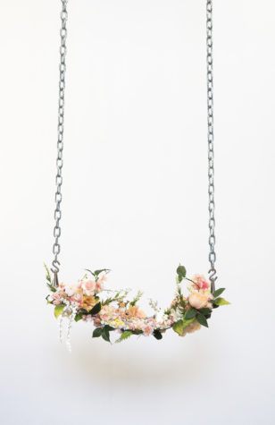 A sculptural artwork depicting a swing with aluminum chain suspension and a seat of peach, pink, white and yellow flowers. The background is white.