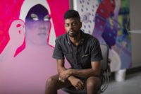 A man sits on a chair, leaning forward, and looking at the camera. There are large, colorful artworks in the background.