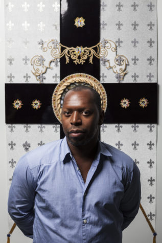A man standing in front of a wall with white wallpaper with silver crosses on it. Behind him is a large, black gross with small gold designs and a round gold design is immediately behind his head. He is looking at the camera and is wearing a blue, button-down shirt.