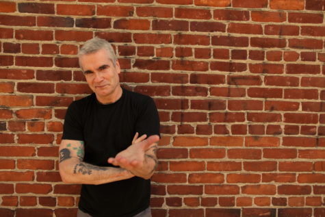 A man stands in front of a red, brick wall, looking at the camera with his left hand out, palm up, towards the camera. He has short, gray hair, tattoos on his arms, and is wearing a black t-shirt.